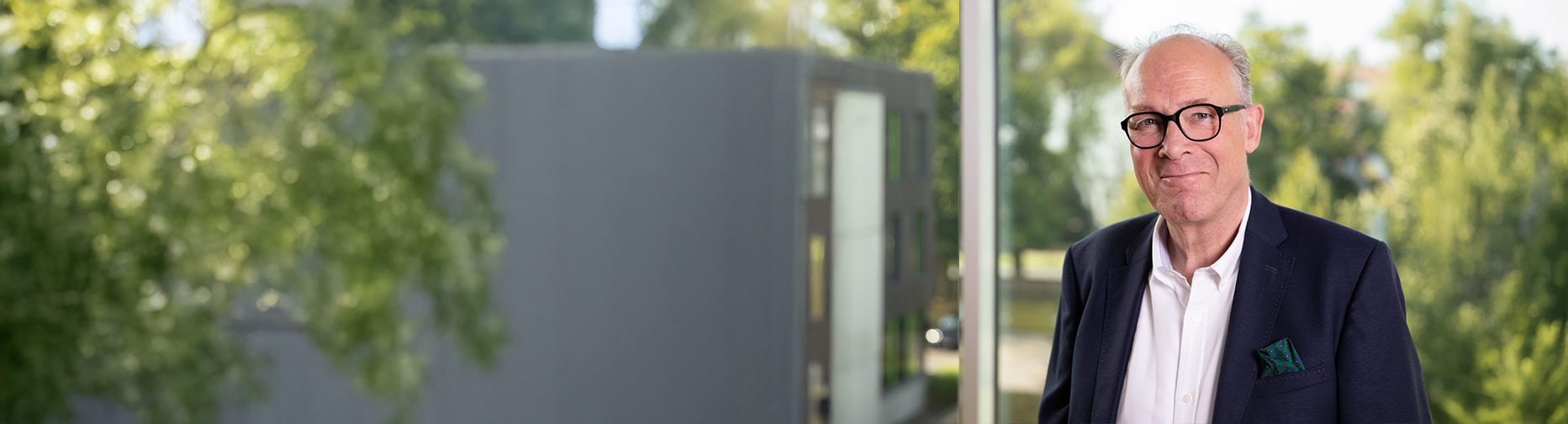 Ein Mann in Anzug und Brille steht vor einem Fenster mit unscharfem Blick auf Bäume und ein modernes Gebäude.