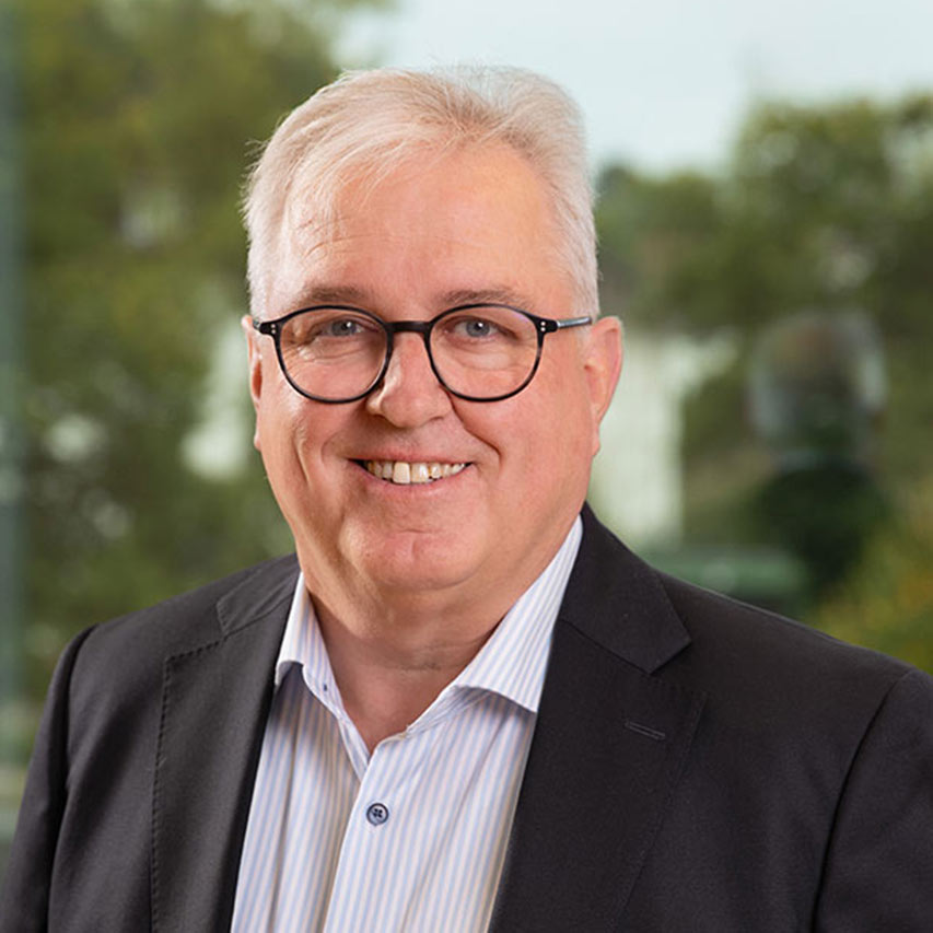 Mann mit grauen Haaren und Brille in Anzug vor verschwommener, grüner Kulisse.