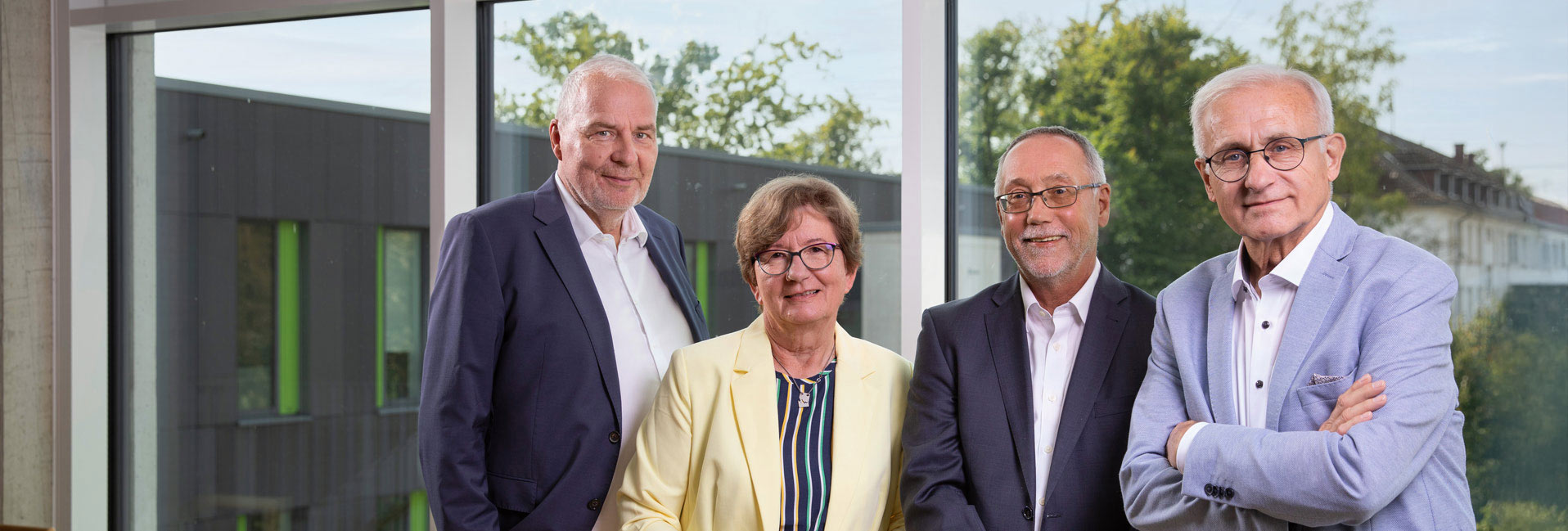 Vier Personen in formeller Kleidung vor einem Fenster, durch das Bäume und Gebäude sichtbar sind.