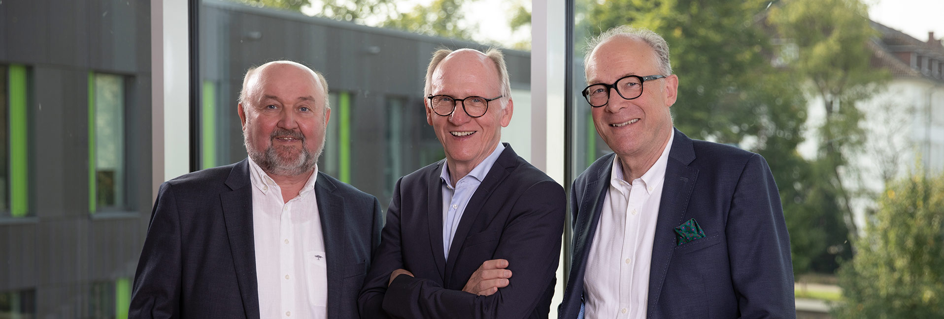 Drei Männer in Anzügen stehen lächelnd vor einer Glasfassade, Bäume im Hintergrund.