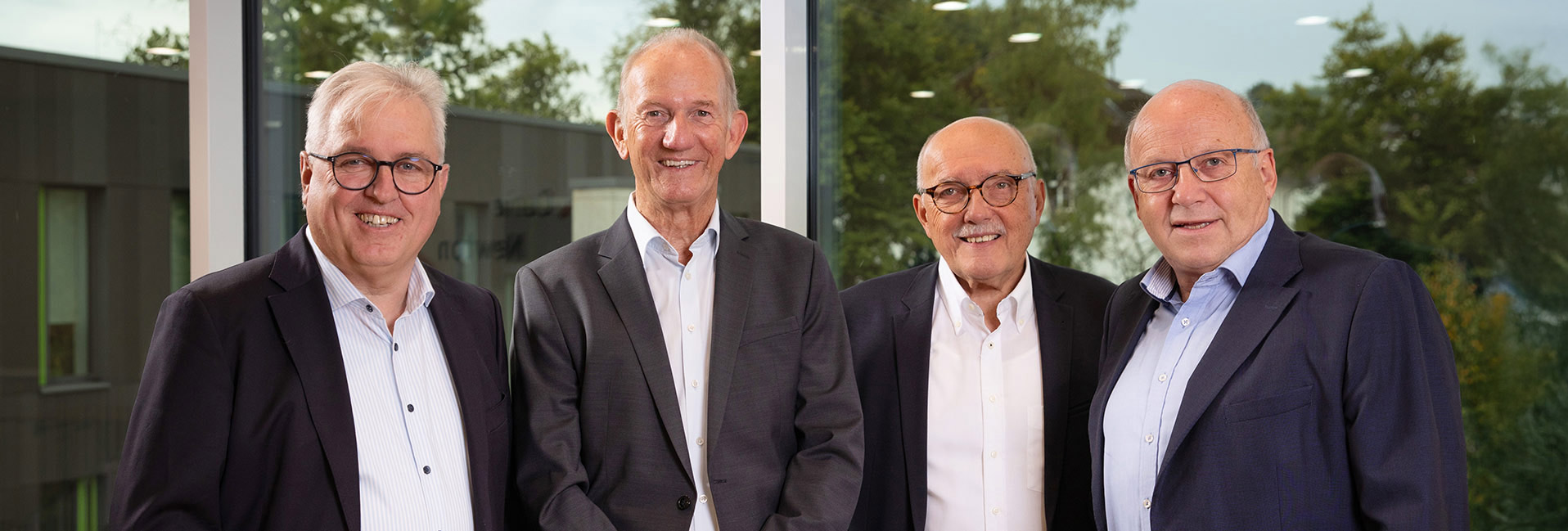 Vier Männer in Anzügen lächeln in die Kamera, stehen vor einem großen Fenster mit Bäumen im Hintergrund.