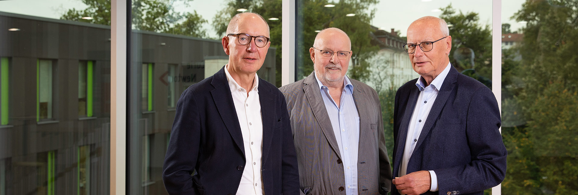 Drei Männer in Anzügen stehen vor großen Fenstern mit Blick auf Bäume und ein Gebäude.