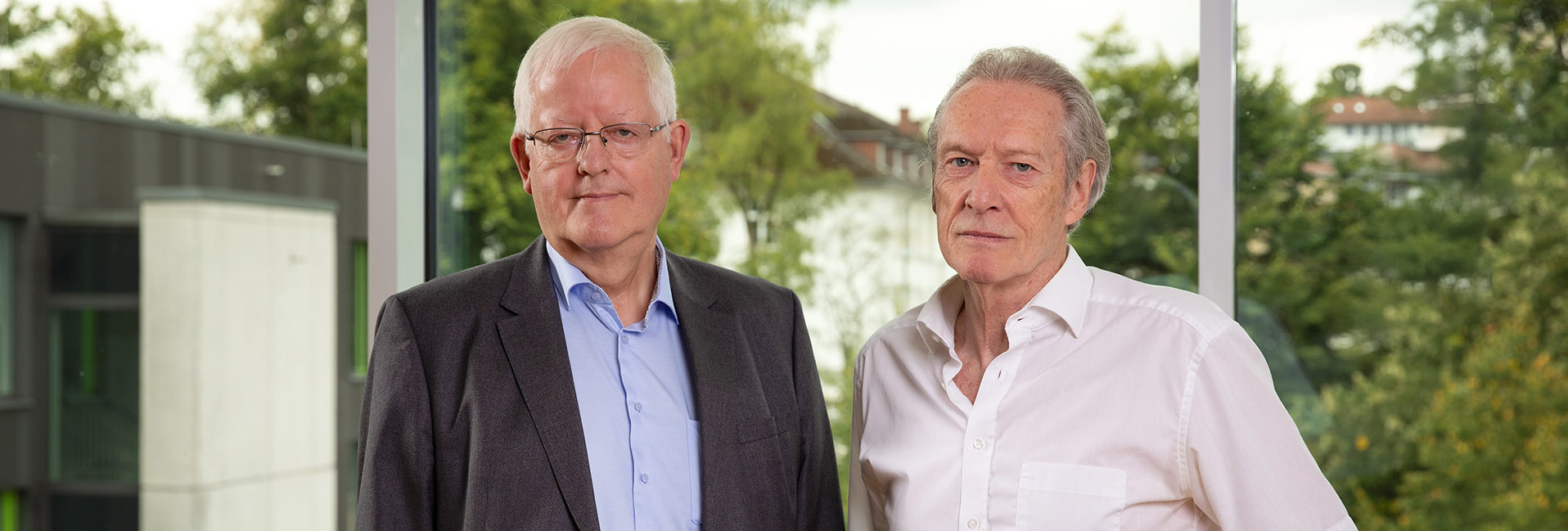 Zwei Männer in Hemd und Anzug stehen vor einem Fenster mit unscharfem Gartenhintergrund.