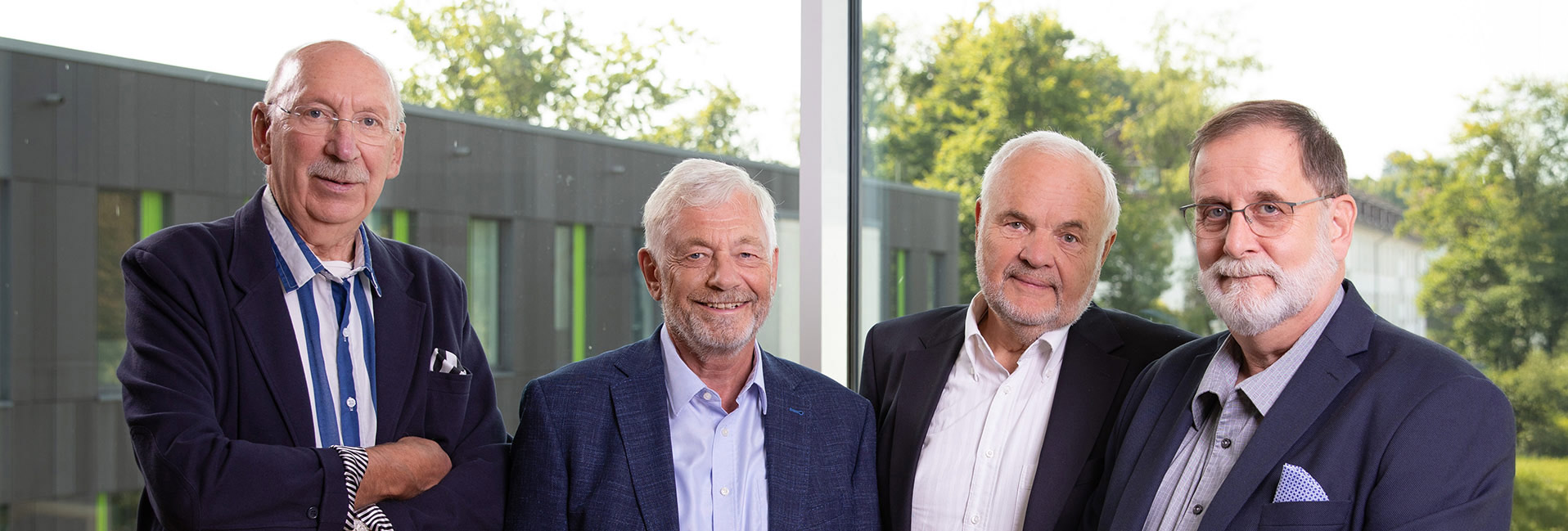 Vier ältere Männer in Anzügen lächeln in die Kamera, vor einem modernen Gebäude mit Glasfront.