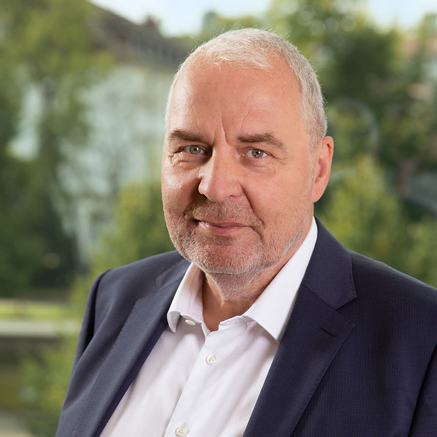 Mann in Anzug vor verschwommenem, grünem Hintergrund, lächelt leicht.