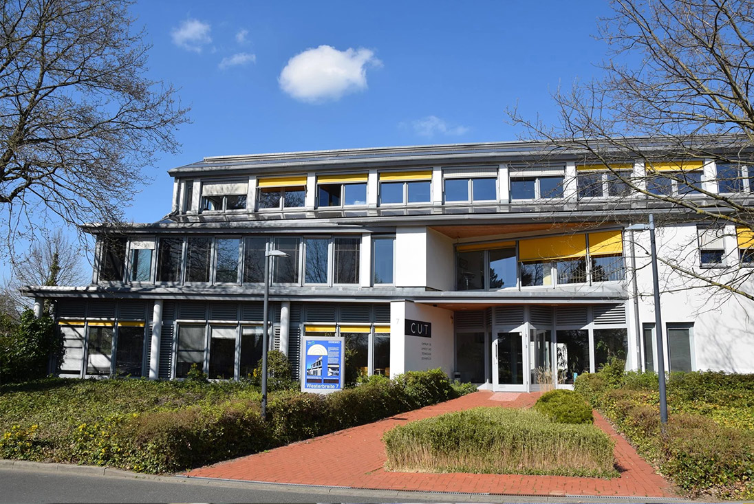 Modernes Gebäude mit gelben Markisen, Glasfronten, rotem Ziegelweg und Garten, unter blauem Himmel.