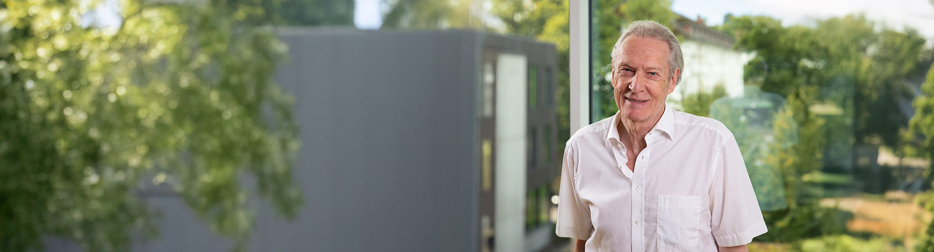 Älterer Mann in weißem Hemd vor einem Fenster mit unscharfem Blick auf Bäume und ein Gebäude.