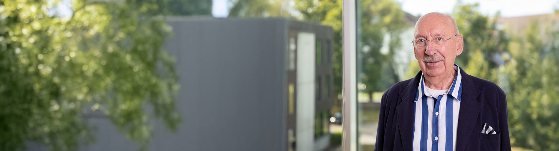 Ein älterer Mann mit Brille und gestreiftem Hemd steht draußen vor einem unscharfen, grünen Hintergrund.