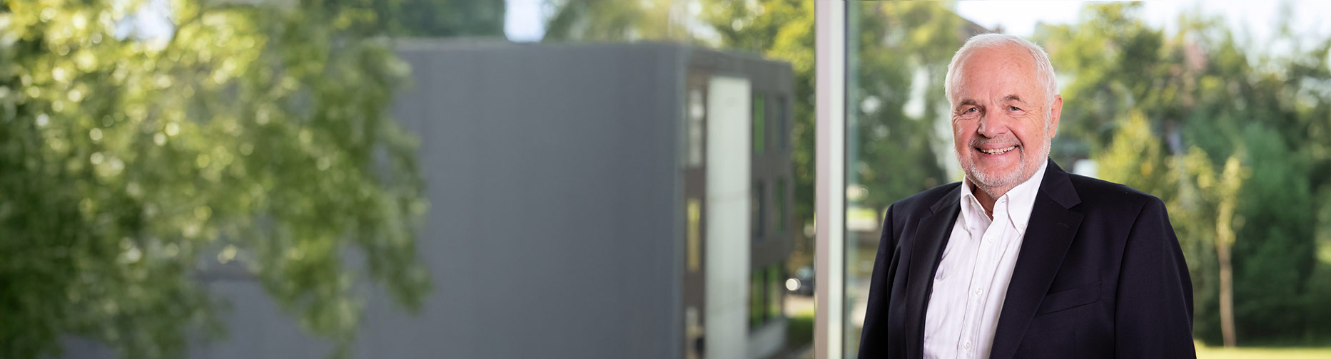 Älterer Mann in Anzug vor Fenster mit Blick auf verschwommene Natur und modernes Gebäude.
