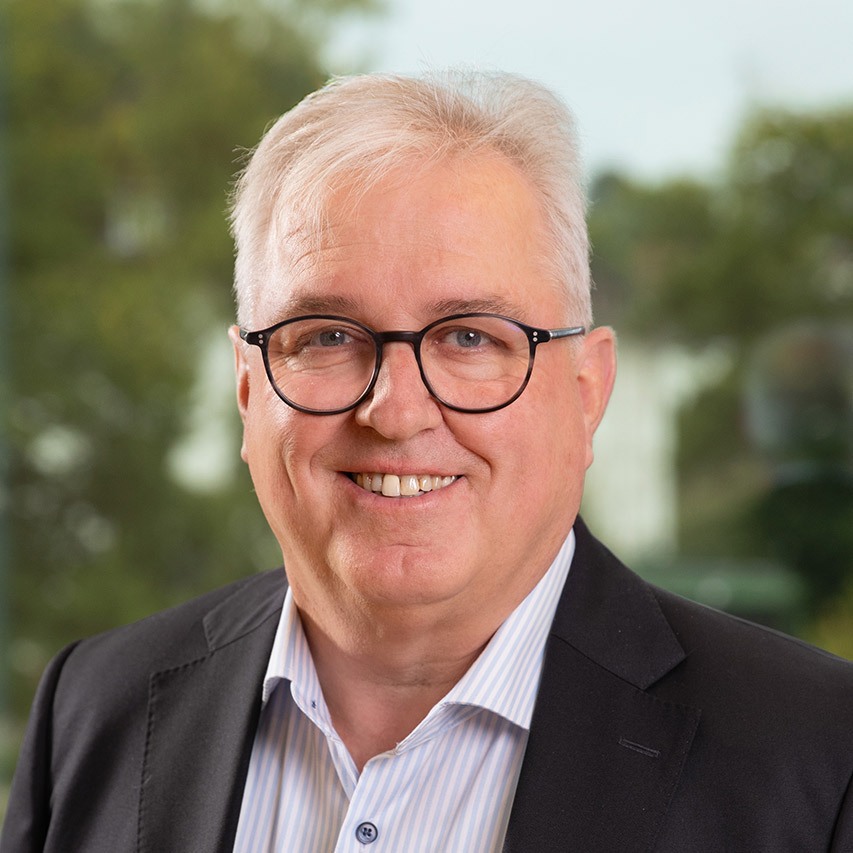 Älterer Mann mit Brille lächelt, trägt dunklen Anzug und gestreiftes Hemd, Hintergrund unscharf mit Bäumen.