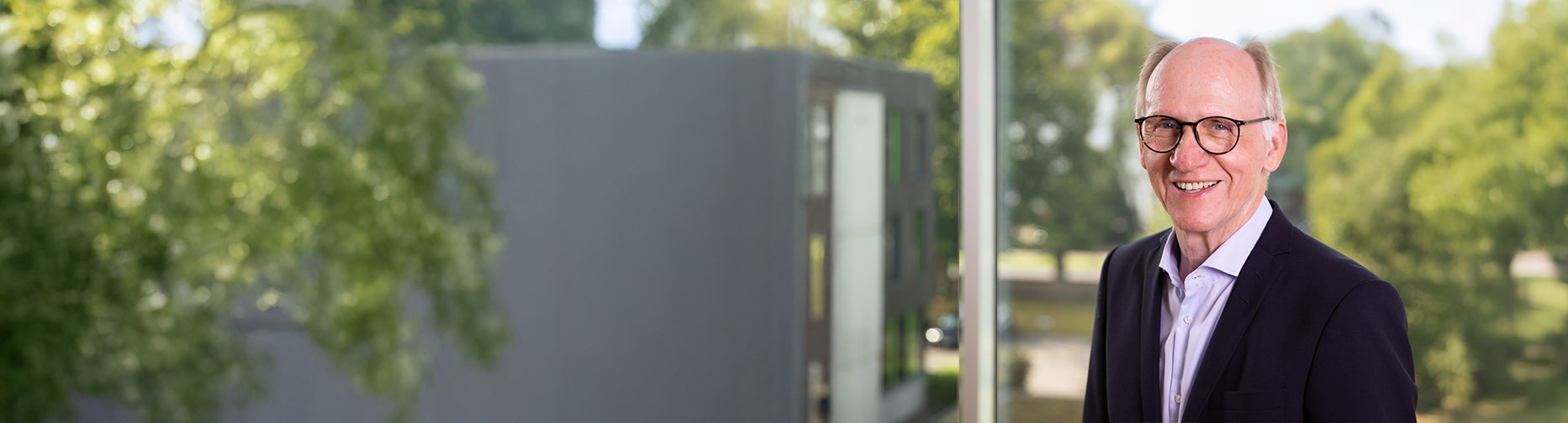 Älterer Mann mit Brille lächelt, steht vor unscharfem grünen Hintergrund und einem modernen Gebäude.