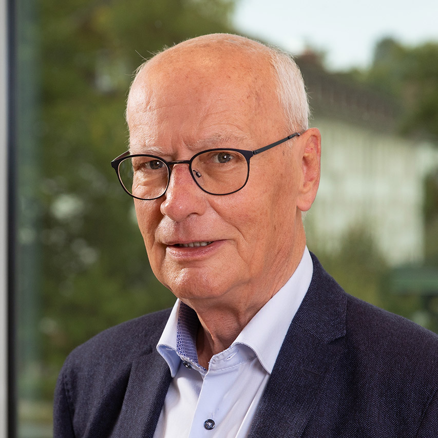 Älterer Mann mit Brille und Anzug, lächelt leicht vor unscharfem Hintergrund mit Bäumen und Gebäuden.