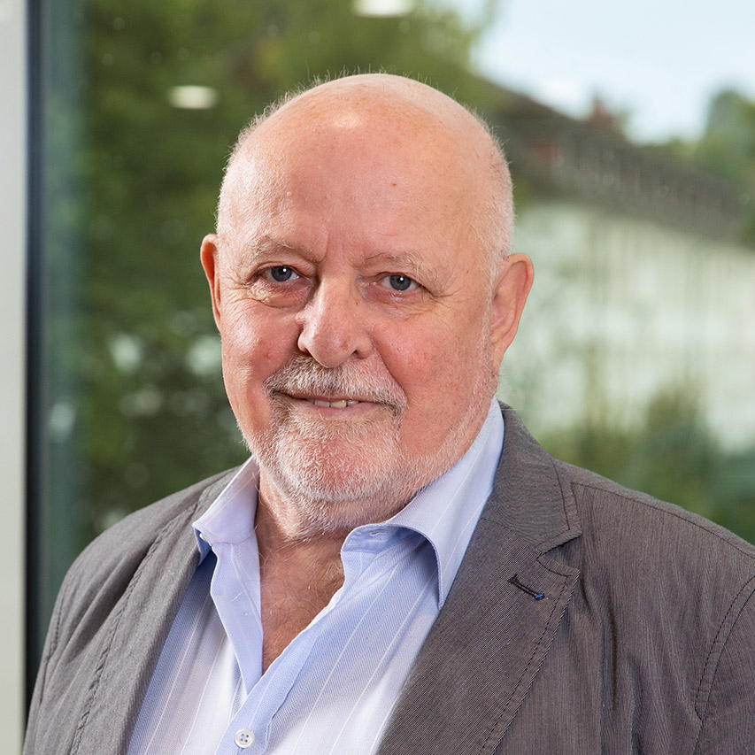 Älterer Mann mit Glatze in grauem Sakko, steht vor Glasfenster mit unscharfem Hintergrund.