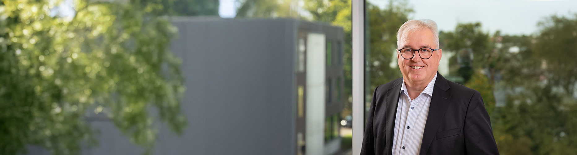 Mann in Anzug mit Brille lächelt vor einem Fenster, draußen Bäume und modernes Gebäude.