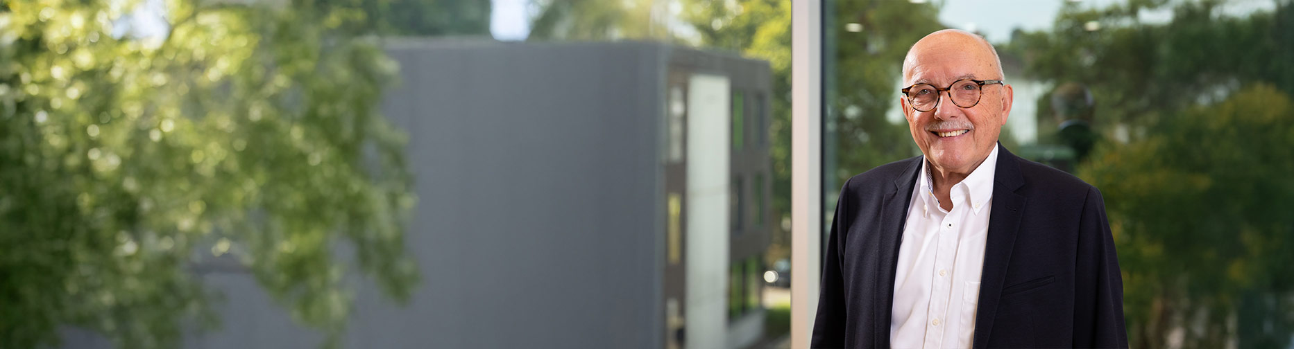 Ein lächelnder Mann mit Brille und Anzug steht vor einem Fenster mit unscharfem Blick auf Bäume und Gebäude.