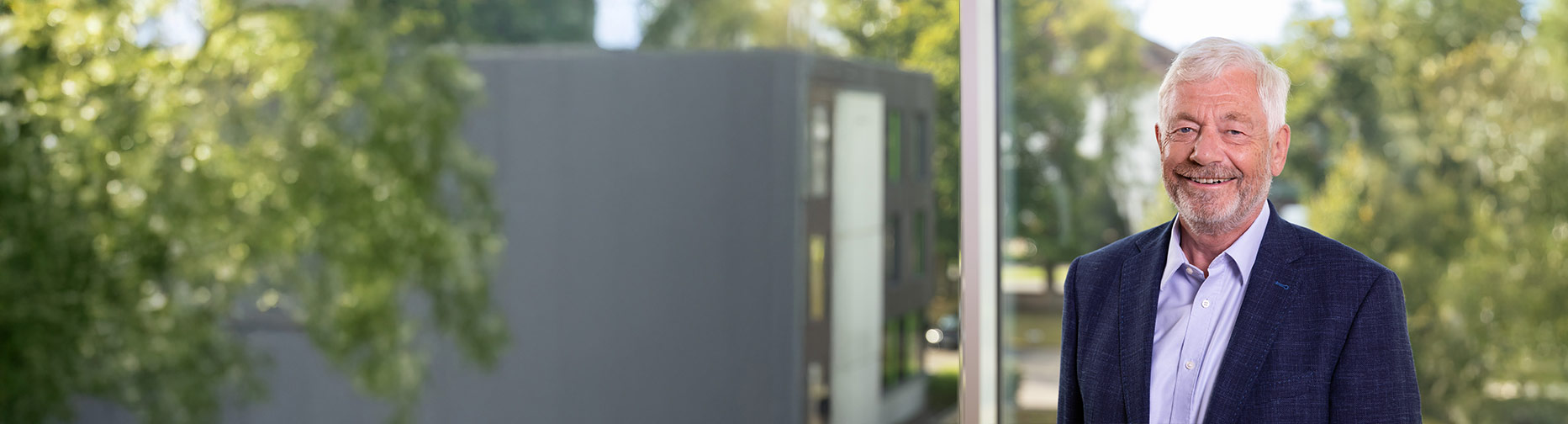 Ein lächelnder Mann in Anzug vor einem Fenster, im Hintergrund verschwommene Bäume und ein graues Gebäude.